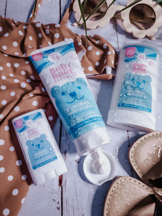 three kids sun care sunscreen products by Baby Bare laid out in a product display with girl's swimsuit sunglasses and sandals 